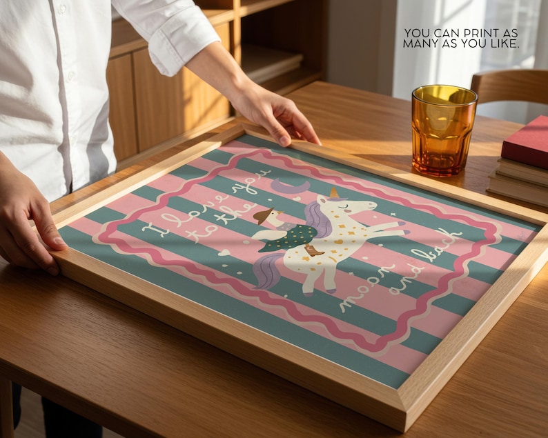 May include: A framed print featuring a whimsical illustration of a unicorn with a rider, set against a striped background in pink and teal. The print includes the text "I love you to the moon and back". The frame is made of light-colored wood.