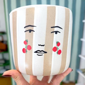 May include: A ceramic planter with a whimsical face design. The planter features vertical stripes in beige and white. The face has black eyebrows, a small mouth, and red berry-like accents on the cheeks. The planter is held by a hand.
