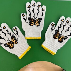 May include: Three white gardening gloves with yellow trim. Each glove features a black butterfly with yellow wings and black floral stems with yellow flowers.