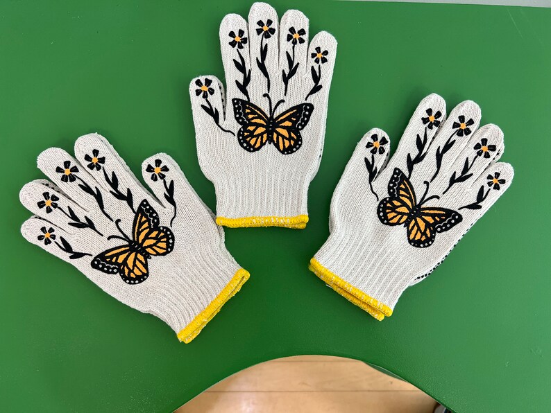 May include: Three white gardening gloves with yellow trim. Each glove features a black butterfly with yellow wings and black floral stems with yellow flowers.