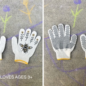 Puede incluir: Un par de guantes de trabajo de algod&oacute;n blanco con ribete amarillo. Un guante tiene un dise&ntilde;o de abeja y flores, el otro tiene puntos negros para agarre. El texto "KIDS GLOVES AGES 3+" est&aacute; impreso debajo de los guantes.