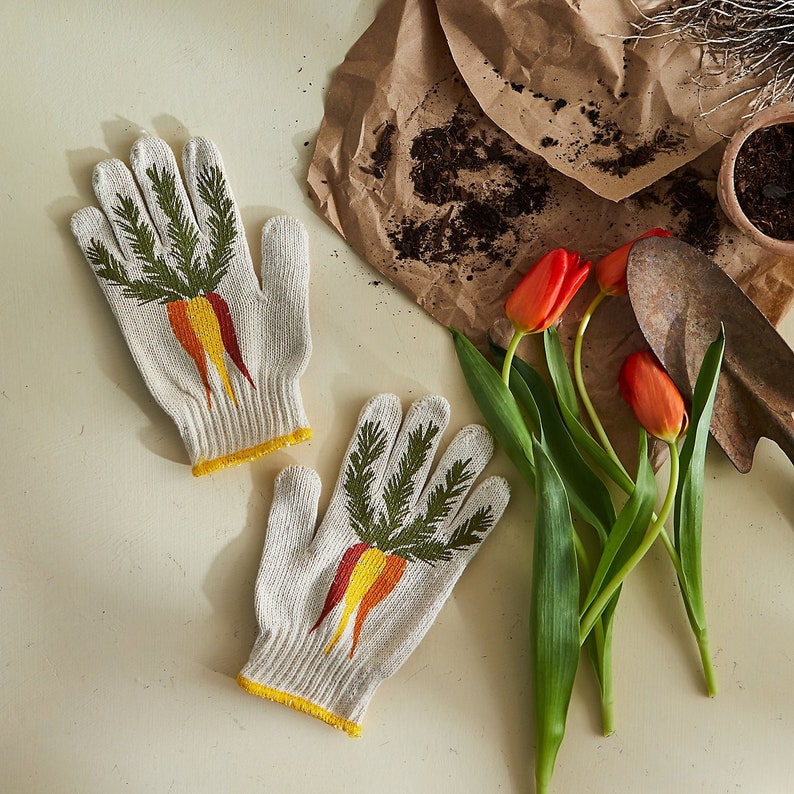 K&ouml;nnte beinhalten: Ein Paar wei&szlig;e Gartenhandschuhe mit gelbem Saum und einem Karotten-Design. Die Handschuhe liegen auf einer wei&szlig;en Oberfl&auml;che mit einem Strau&szlig; orangefarbener Tulpen und einer braunen Papiert&uuml;te.