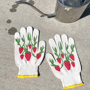 May include: A pair of white gardening gloves with a yellow trim and a strawberry print. The gloves are on a gray concrete surface.