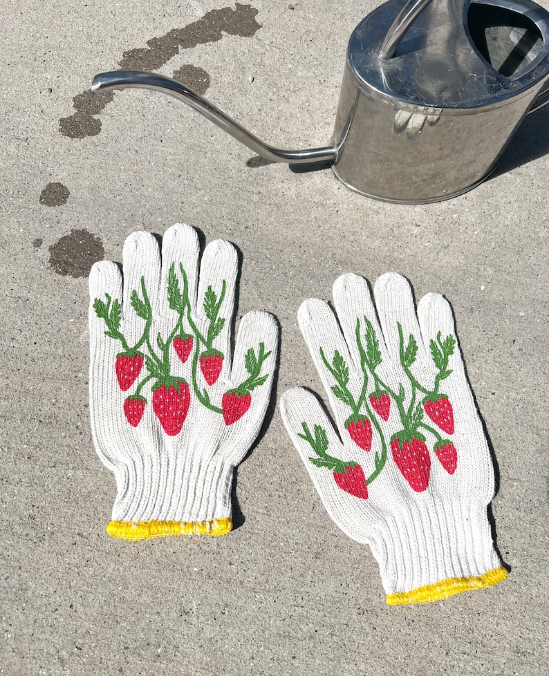 May include: A pair of white gardening gloves with a yellow trim and a strawberry print. The gloves are on a gray concrete surface.