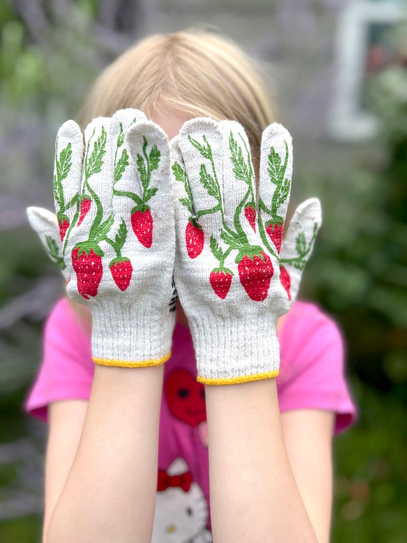 Puede incluir: Un par de guantes de jardiner&iacute;a de algod&oacute;n blanco con estampado de fresas. Los guantes tienen un ribete amarillo alrededor de las mu&ntilde;ecas.