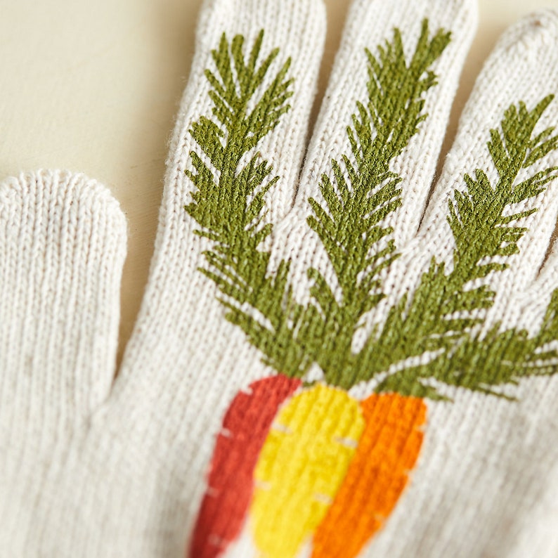 K&ouml;nnte beinhalten: Ein Paar wei&szlig;e Strickhandschuhe mit einem gr&uuml;nen Karotten-Design. Die Karotte hat orange, gelbe und rote Akzente.