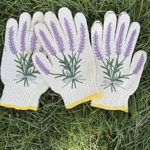 May include: A set of three white gardening gloves with a lavender floral design. The gloves feature a purple lavender sprig with green leaves and a yellow trim. The gloves are lying on a bed of green grass.