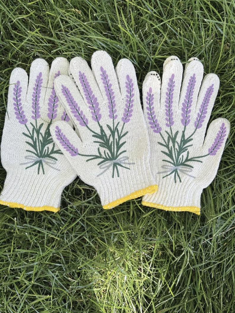 May include: A set of three white gardening gloves with a lavender floral design. The gloves feature a purple lavender sprig with green leaves and a yellow trim. The gloves are lying on a bed of green grass.