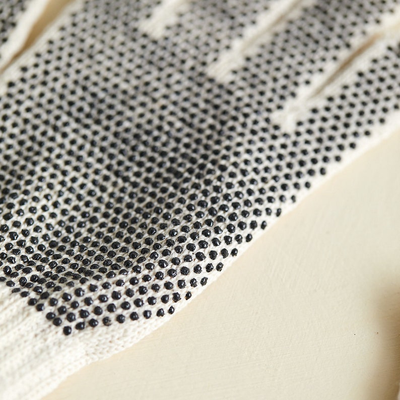 K&ouml;nnte beinhalten: Nahaufnahme eines wei&szlig;en Baumwollhandschuhs mit schwarzen Punkten an den Fingerspitzen. Der Handschuh ist aus einem weichen, atmungsaktiven Material gefertigt und f&uuml;r Grip und Fingerfertigkeit konzipiert.
