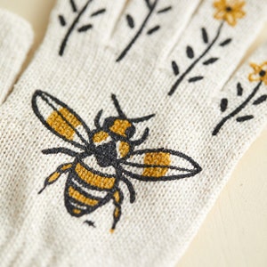 Puede incluir: Un par de guantes de punto blanco con un dise&ntilde;o de abeja negra y amarilla. La abeja est&aacute; bordada en el guante y tiene un dise&ntilde;o detallado.