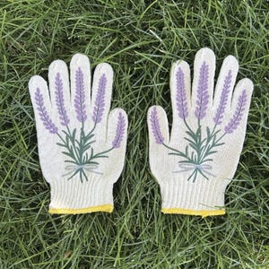 May include: A pair of gardening gloves with a lavender design. The gloves are off-white with a yellow trim at the wrist. Lavender sprigs are printed on the back of each glove in purple and green.