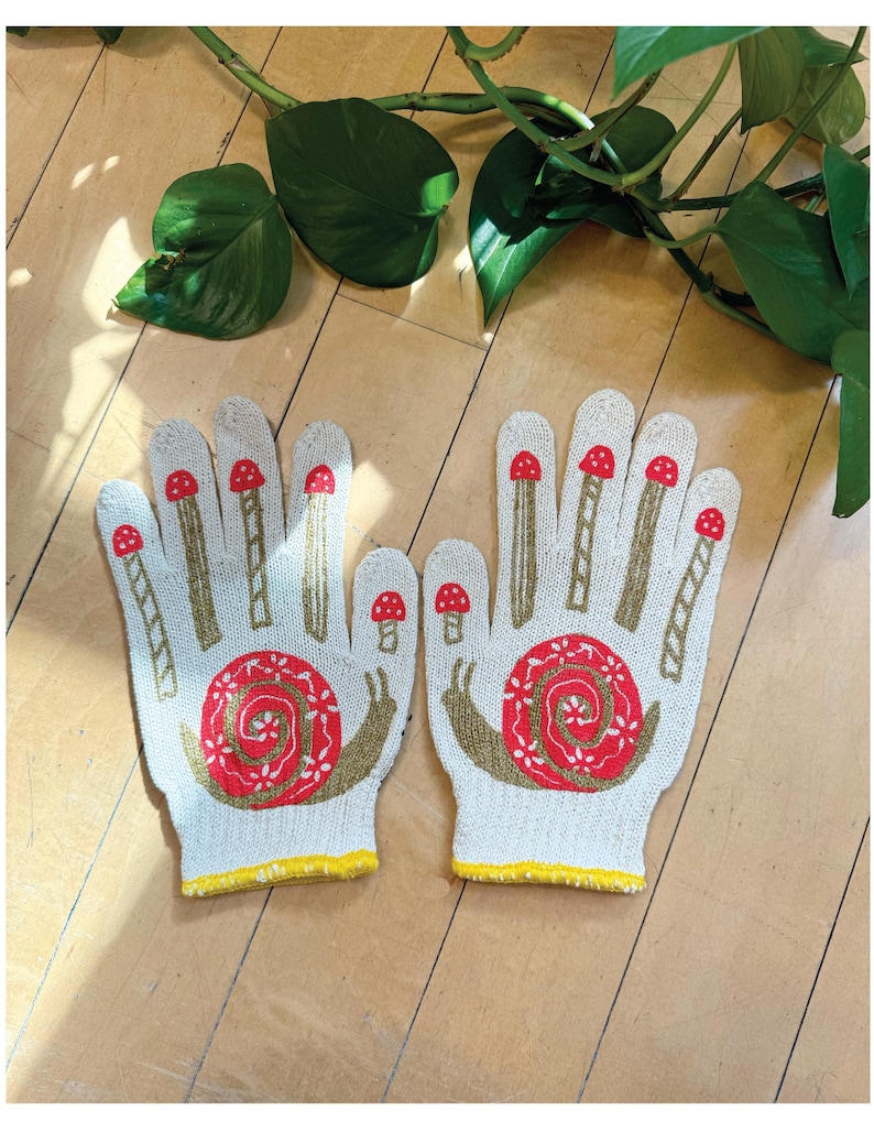 May include: A pair of white gardening gloves with a yellow trim. The gloves feature a red and brown snail design with mushrooms on the fingers.