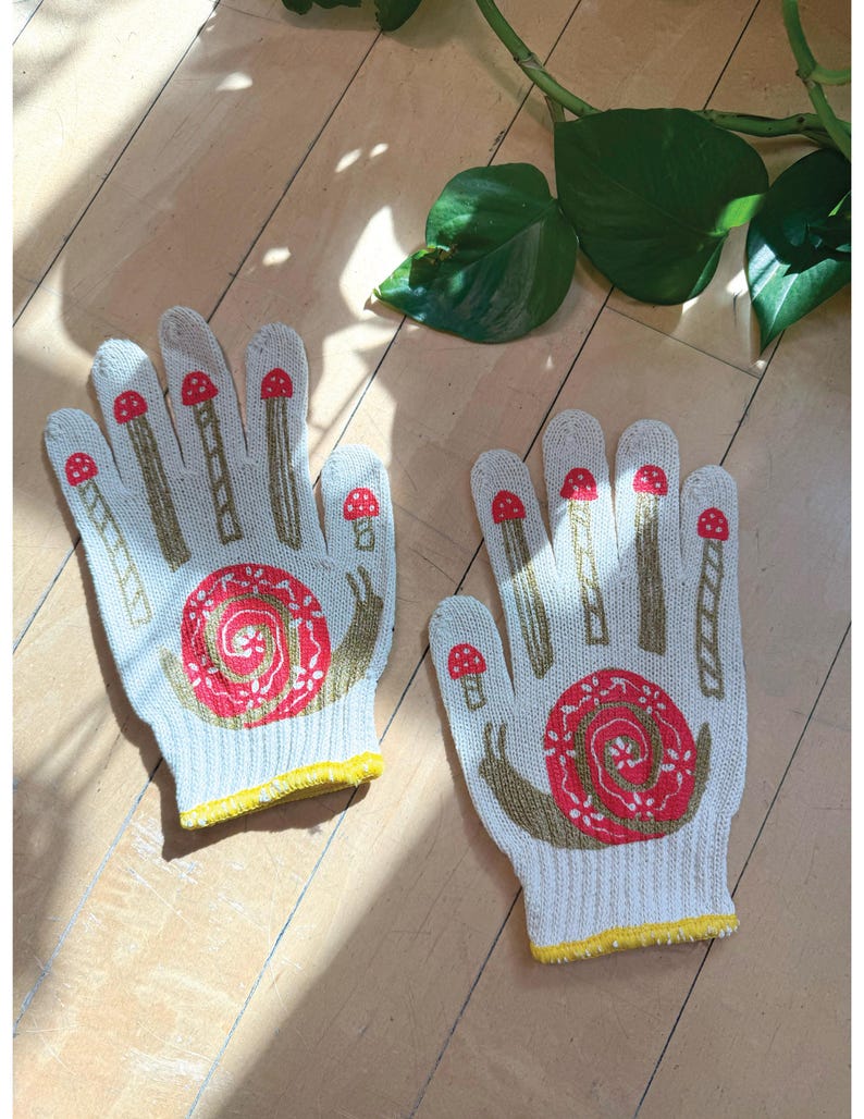May include: A pair of white gardening gloves with a yellow trim. The gloves feature a red and brown snail design with mushrooms on the fingers.