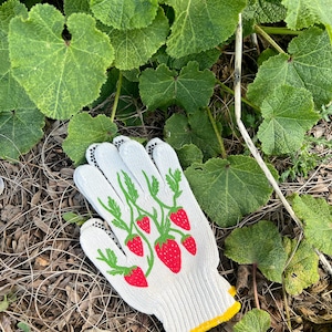 May include: White gardening gloves with a pattern of red strawberries and green leaves on the back of the hand. The gloves are lying on a bed of brown mulch and green leaves.