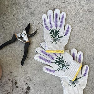 May include: White gardening gloves with lavender sprig embroidery and yellow trim are paired with black and silver pruning shears. The gloves feature purple lavender and green leaves, perfect for gardening.