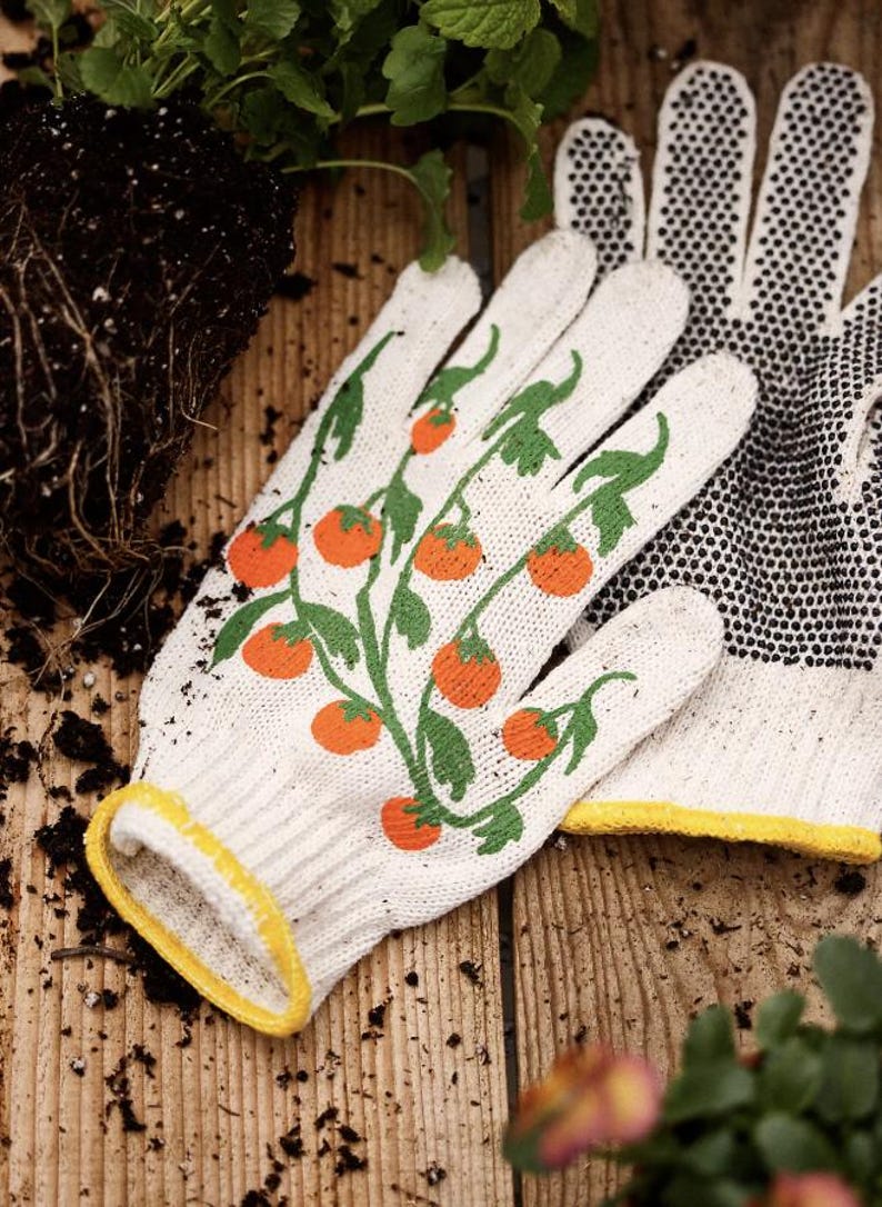 May include: White gardening gloves with a tomato plant design. The gloves have a yellow trim and a dotted pattern on the palm. The design features orange tomatoes and green leaves.