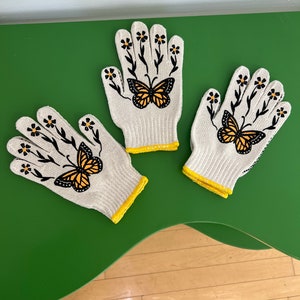May include: Three pairs of white gardening gloves with yellow trim. Each glove has a black butterfly and floral design.