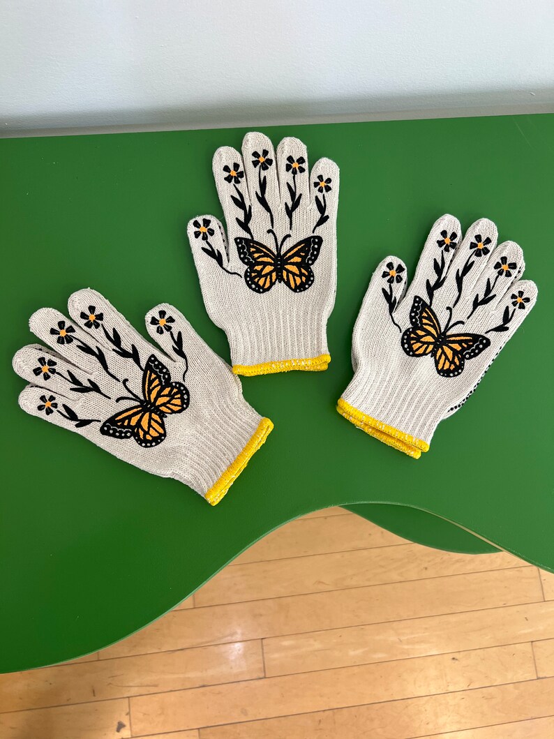 May include: Three pairs of white gardening gloves with yellow trim. Each glove has a black butterfly and floral design.
