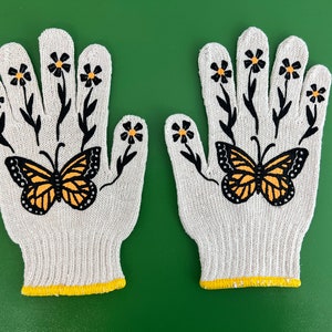 May include: A pair of white gardening gloves with a yellow trim. The gloves are decorated with a black butterfly and yellow flowers.