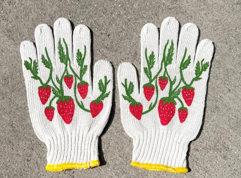 Puede incluir: Un par de guantes de jardiner&iacute;a de algod&oacute;n blanco con ribete amarillo y dise&ntilde;o de fresas. Los guantes tienen un patr&oacute;n de fresas rojas y verdes en la parte posterior de la mano.
