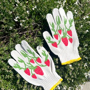 May include: A pair of white gardening gloves with a yellow trim. The gloves have a pattern of green vines and red strawberries.