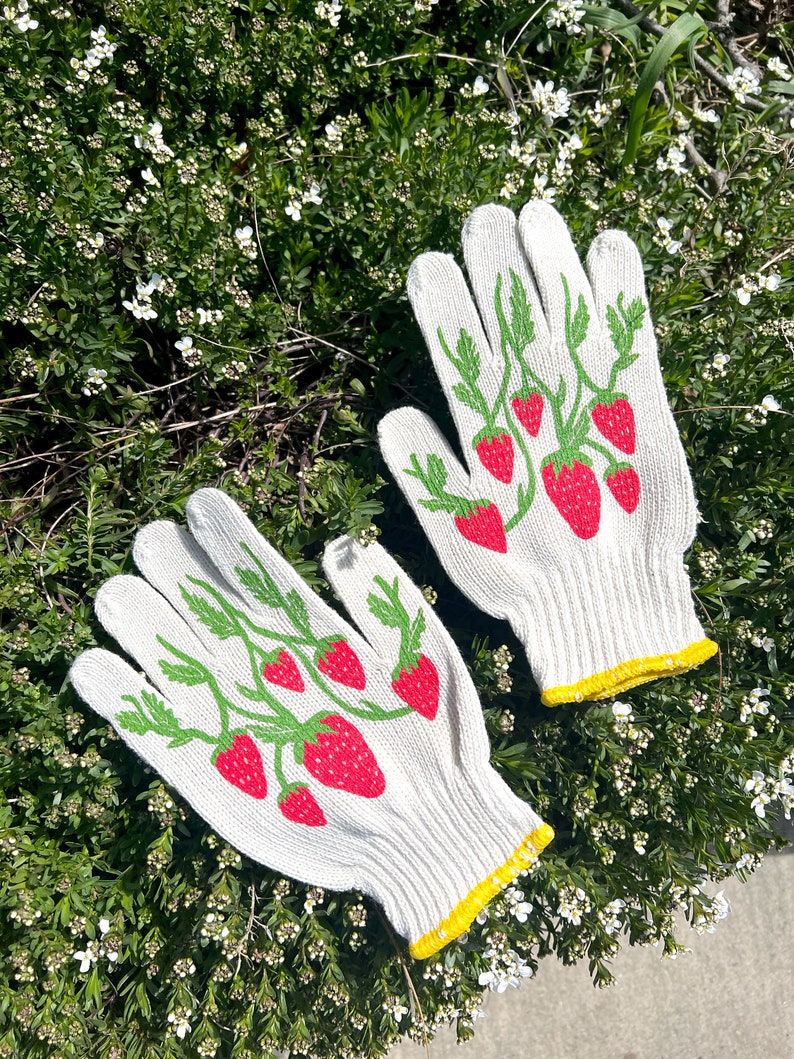 May include: A pair of white gardening gloves with a yellow trim. The gloves have a pattern of green vines and red strawberries.