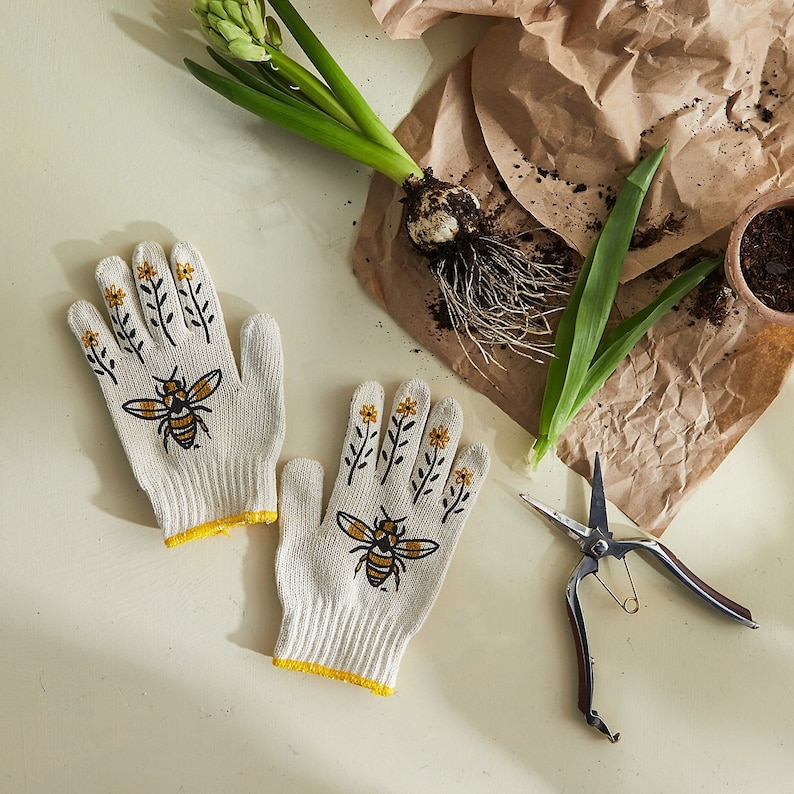 Puede incluir: Un par de guantes de jardiner&iacute;a blancos con ribete amarillo y un dise&ntilde;o de abeja y flores. Los guantes est&aacute;n sobre una superficie blanca con unas tijeras de jard&iacute;n.