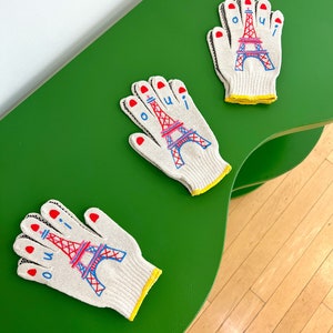 May include: Three white work gloves with red, blue, and pink embroidery of the Eiffel Tower and the word "oui". The gloves are on a green surface.