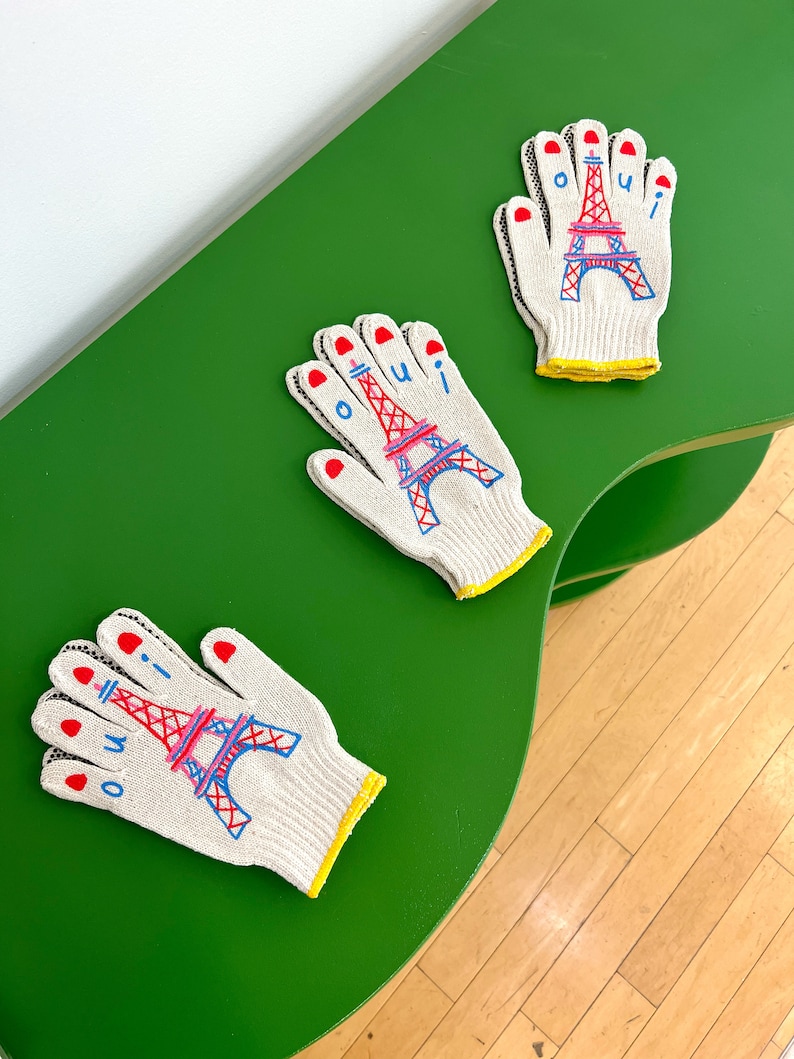 May include: Three white work gloves with red, blue, and pink embroidery of the Eiffel Tower and the word "oui". The gloves are on a green surface.