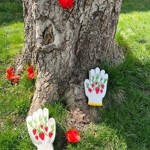 May include: Two white gardening gloves with a strawberry design on a green background. The gloves are lying on the ground near a tree trunk and red flowers.