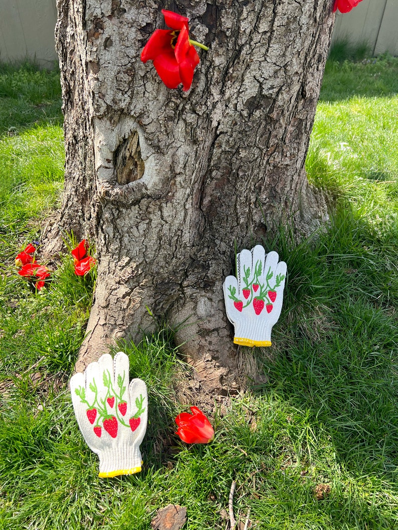 May include: Two white gardening gloves with a strawberry design on a green background. The gloves are lying on the ground near a tree trunk and red flowers.