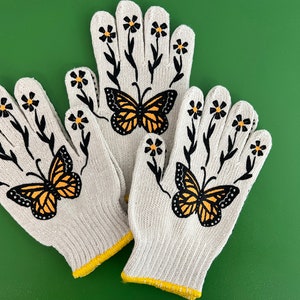 May include: A pair of white gardening gloves with black floral designs and orange butterflies. The gloves have yellow trim around the wrists.