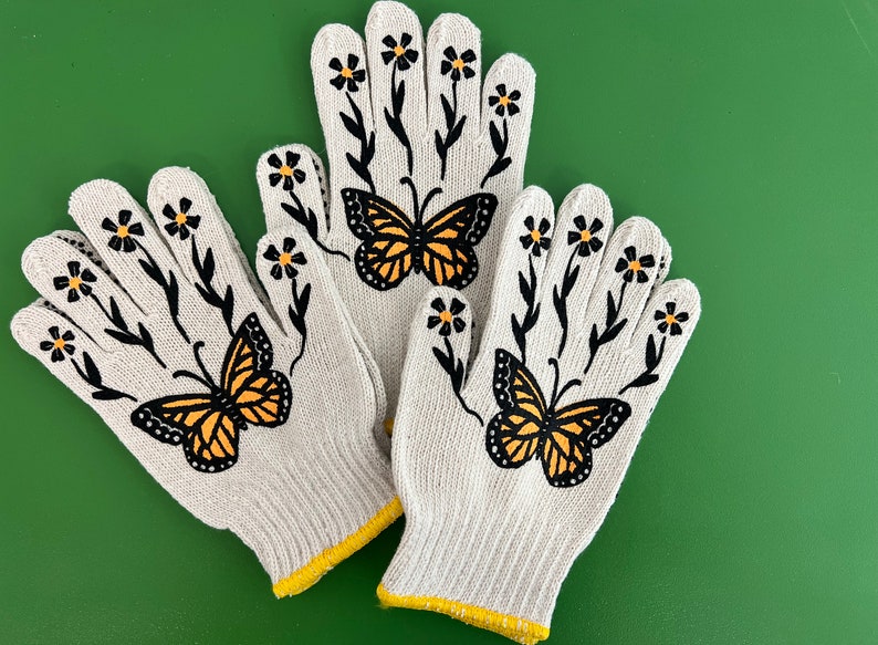 May include: A pair of white gardening gloves with black floral designs and orange butterflies. The gloves have yellow trim around the wrists.