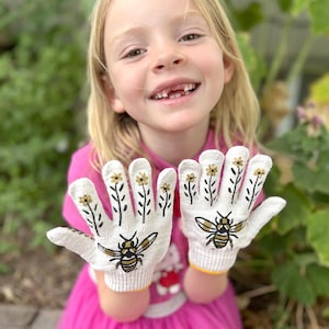 May include: White knit gloves with a bee and flower design. The gloves feature a yellow cuff and black and gold illustrations of bees and flowers. The gloves are being held up by a person wearing a pink shirt.