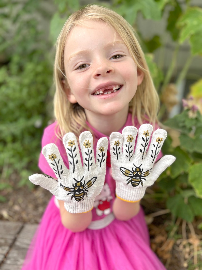 May include: White knit gloves with a bee and flower design. The gloves feature a yellow cuff and black and gold illustrations of bees and flowers. The gloves are being held up by a person wearing a pink shirt.