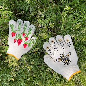 Puede incluir: Un par de guantes de jardiner&iacute;a blancos con ribete amarillo. Un guante tiene un dise&ntilde;o de fresa y el otro un dise&ntilde;o de abeja.
