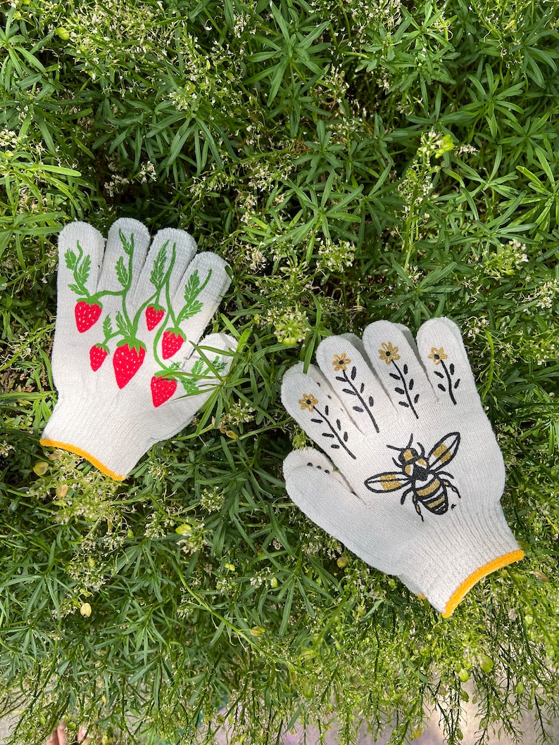 Puede incluir: Un par de guantes de jardiner&iacute;a blancos con ribete amarillo. Un guante tiene un dise&ntilde;o de fresa y el otro un dise&ntilde;o de abeja.