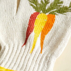 K&ouml;nnte beinhalten: Ein wei&szlig;er gestrickter Gartenhandschuh mit einem bunten Karotten-Design. Der Handschuh hat ein gelbes, orangefarbenes und rotes Karotten-Design.