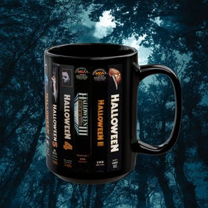 Puede incluir: Taza de café negra con un diseño de cintas VHS para las películas de Halloween. Las cintas están etiquetadas como "Halloween", "Halloween II", "Halloween III: La Temporada de la Bruja", "Halloween 4" y "Halloween 5".