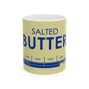 May include: A ceramic mug with a light yellow exterior and a white interior. The mug features the words "SALTED BUTTER" in large blue letters. Below the text are blue lines with measurements in tablespoons and cups.