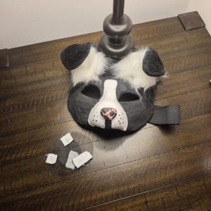 May include: A black and white animal mask featuring faux fur ears, a white snout with a pink and black nose, and eye cutouts. A black elastic strap is attached to the side. A separate small black mesh piece with white adhesive squares is included.