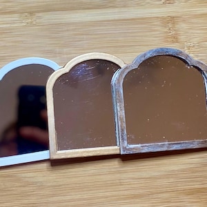 May include: Three arched mirrors with different colored frames. The mirror on the left has a white frame, the middle mirror has a gold frame, and the mirror on the right has a brown frame.