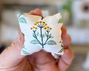 May include: A small, square, off-white fabric pillow featuring a repeating botanical print of yellow flowers with green stems and leaves. The textile has a visible woven texture, and the pattern is centered on the front of the miniature cushion.
