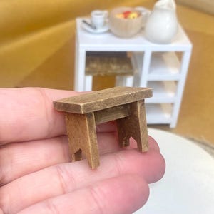 May include: A miniature wooden table with a rectangular top and four legs. The table is a light brown color and appears to be handmade. In the background, a white table with a small stool, a bowl, and a pitcher are visible.