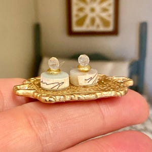 Pode incluir: Duas garrafas cilíndricas em miniatura com bases verde-pálido e tampas transparentes facetadas, cada uma com uma etiqueta de papel branco com escrita preta à mão, sobre uma pequena bandeja retangular de metal dourado ornamentado.