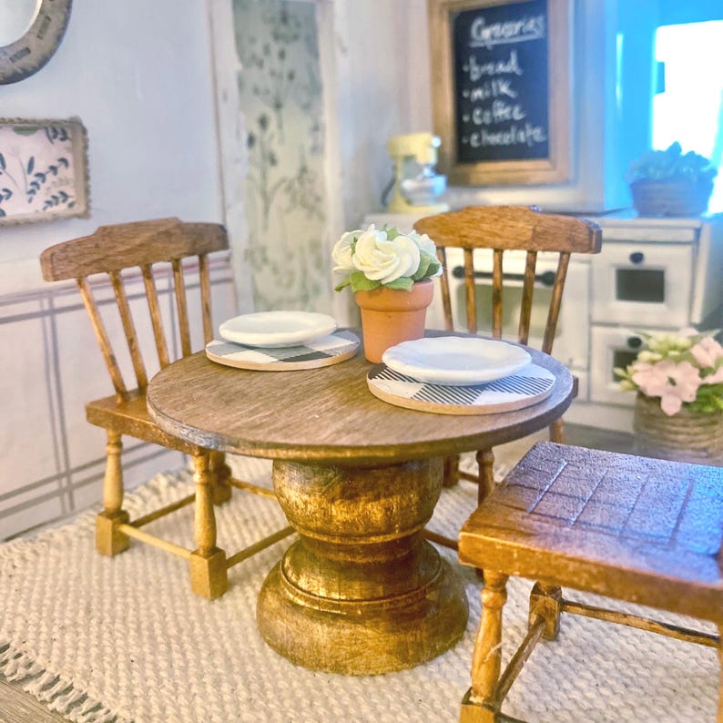 May include: A miniature wooden dining set featuring a round pedestal table with two chairs, two white plates on blue and white checkered placemats, and a small terracotta pot containing white flowers, set in a dollhouse kitchen scene.