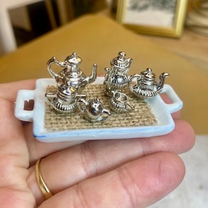 Op de afbeelding: Een zilverkleurige miniatuur theeset bestaande uit een theepot, koffiepot, melkkannetje, suikerpot en een klein kopje op een rechthoekig wit dienblad met een inleg van gestructureerd beige jute.