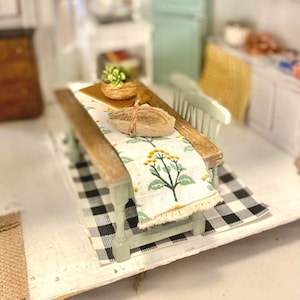 May include: A miniature wooden dining table with light green legs and a natural wood top, covered by a white table runner featuring a green and yellow floral pattern. A small woven basket and a tiny potted plant sit on the table surface.