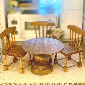 May include: A miniature wooden dining set consisting of a round table with a pedestal base and two matching chairs with spindle backs and square seats, all finished in a medium brown wood stain with visible grain texture.