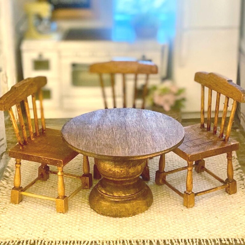 May include: A miniature wooden dining set consisting of a round table with a pedestal base and two matching chairs with spindle backs and square seats, all finished in a medium brown wood stain with visible grain texture.